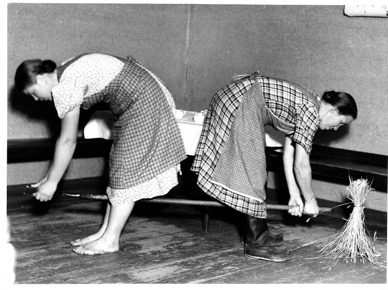 Esikatselukuva:&nbsp;Illanvietto. Eevi Kiiski (vas.) ja Ida Asikainen. Kuvattu Inkisen tuvassa. Kaksi naista seisoo selät vastakkain ja pitää heinähankoa jalkojensa välissä yrittäen kaataa edessään olevan olkilyhdettä.